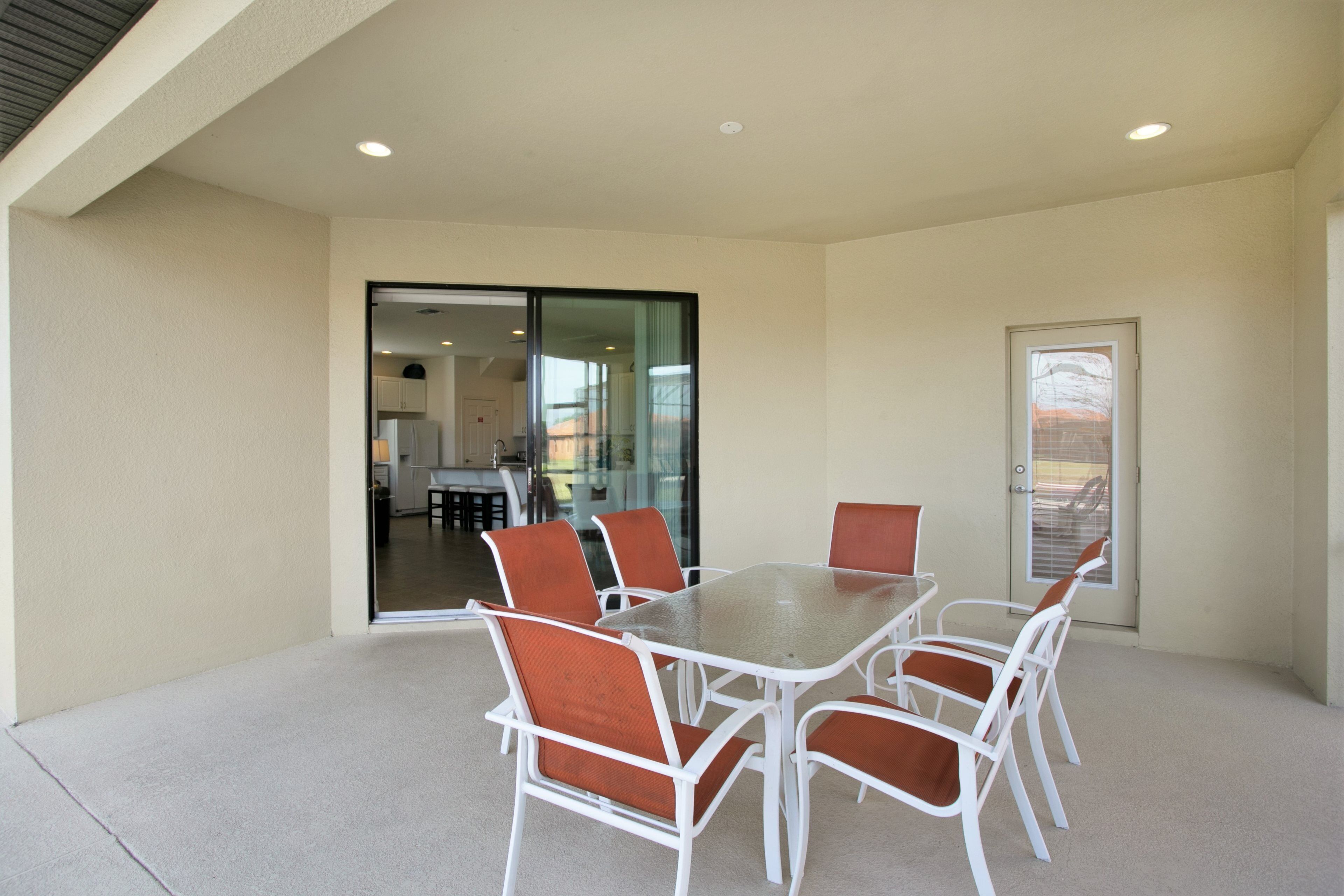 A covered outdoor dining area with a glass table and white chairs, a sliding glass door leading to a kitchen, and a door with a window view.