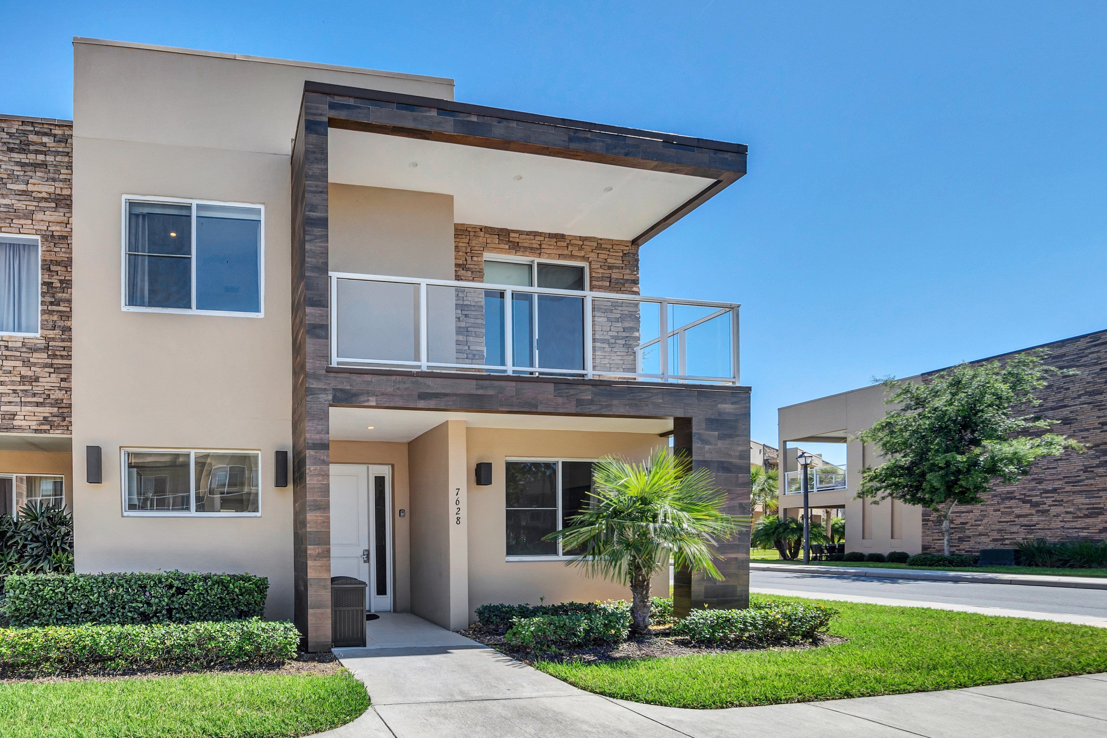 A modern two-story residential building with a balcony, a small front yard, and a clear sky.