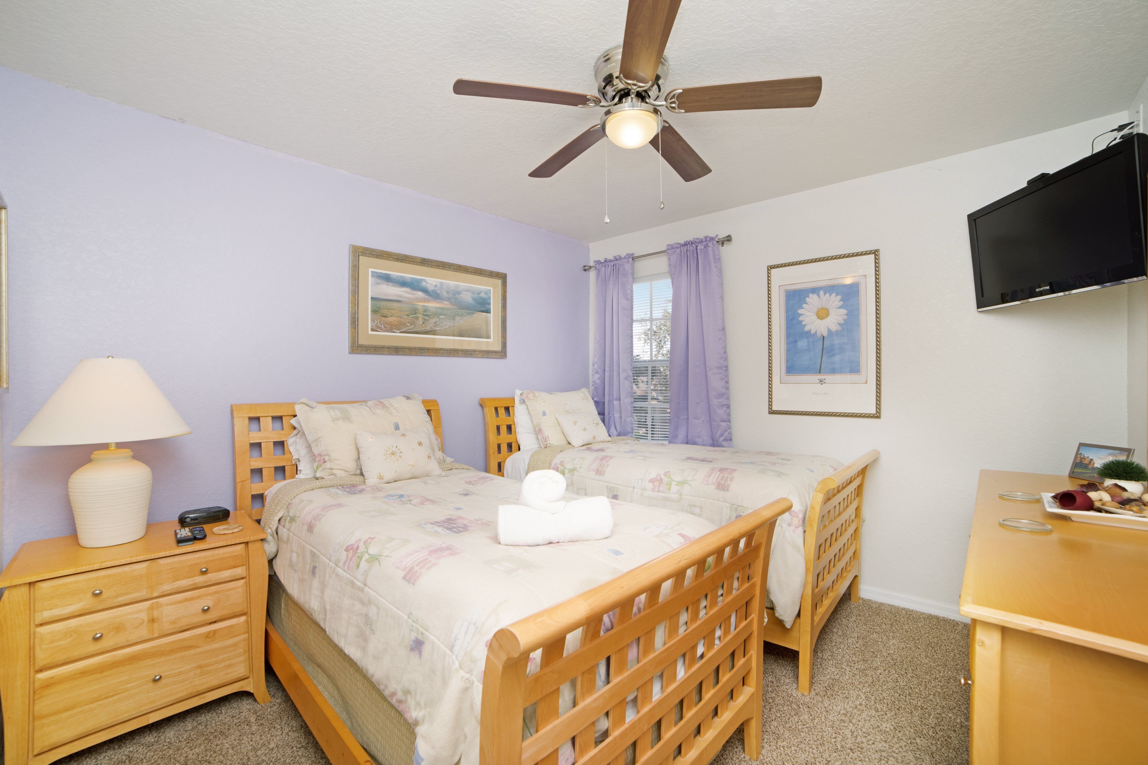 A bedroom with two wooden beds, a ceiling fan, a nightstand with a lamp, and a TV on the wall.