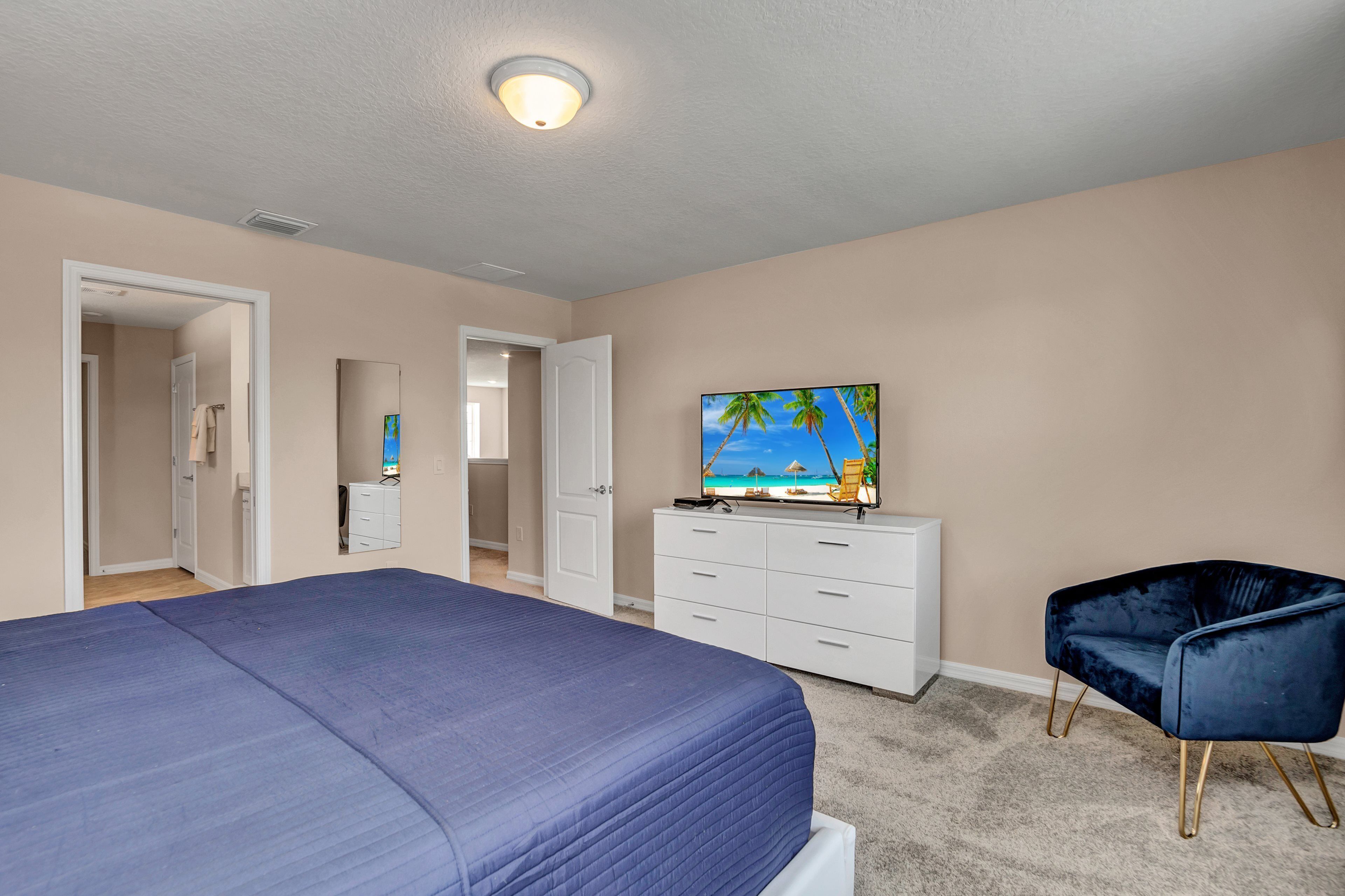 Ein Schlafzimmer mit einem großen Bett, einem Fernseher auf einer Kommode, einem Sessel und einer Tür zu einem anderen Raum.