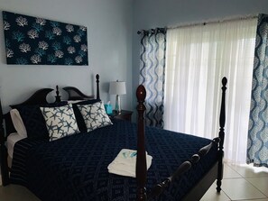 A neatly made bed with a dark blue bedspread and star-patterned pillows, a bedside table with a lamp, and a wall-mounted artwork. - Luxury spacious condo in the heart of it all (Barbados)