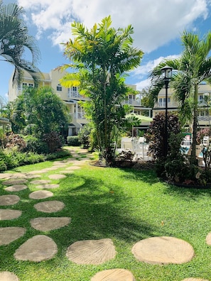 A pathway with circular stepping stones leading to a building with multiple balconies and a well-maintained garden with various plants and a palm tree.