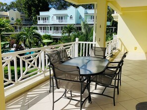 A balcony with a glass table and chairs, surrounded by a white railing, overlooking a pool and buildings. - Luxury spacious condo in the heart of it all (Barbados)