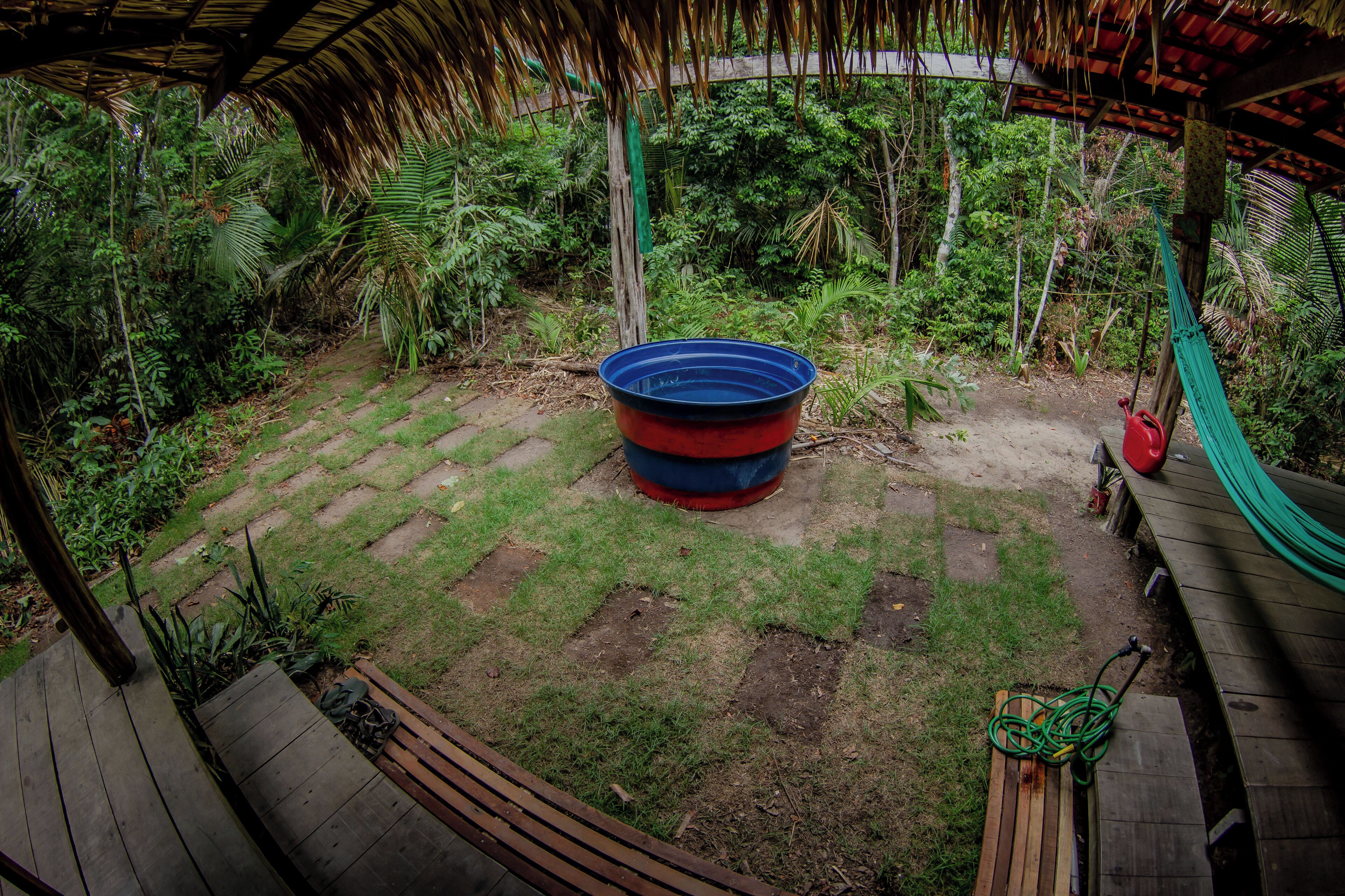 A wooden deck with a hammock and a blue and red container in a garden area with a thatched roof.