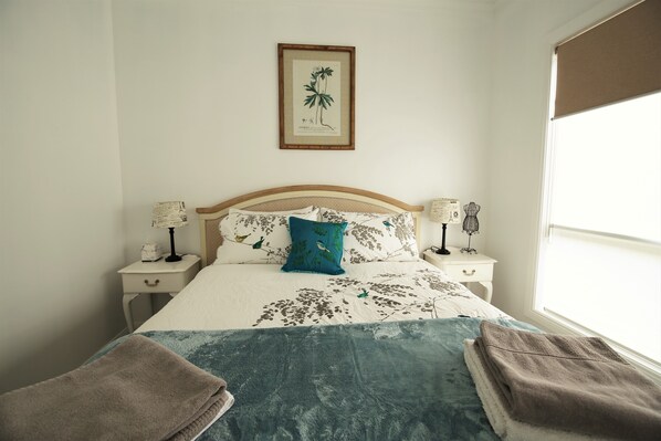 A neatly made bed with floral bedding, two bedside tables with lamps, a framed botanical print on the wall, and a window with a blind.