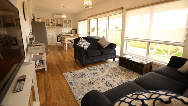 A spacious living room with a dark sofa, a coffee table, and a large window offering a view of the outdoors. - Bridgecroft Villa (Richmond)