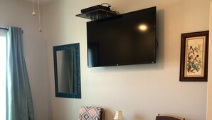 A hotel room with a bed, two chairs, a small table with a lamp, a TV mounted on the wall, and a framed picture.