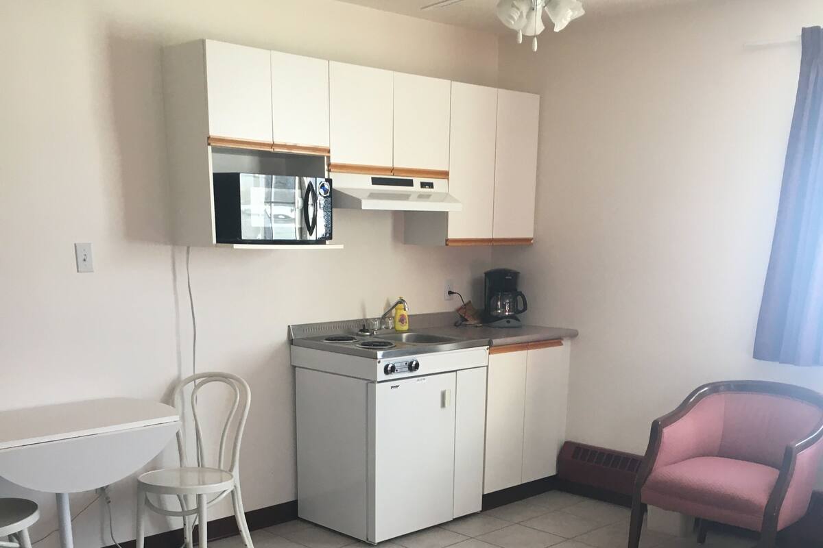 A compact kitchen with a gas stove, white cabinets, a microwave, and a coffee maker. There is a dining table with two chairs and a pink chair.