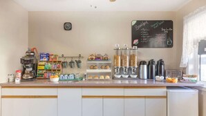 Ein Frühstücksbuffet mit einer Kreidetafel, auf der Speisen wie Muffins, Müsli und Obstsalat aufgeführt sind. Auf der Anrichte befinden sich verschiedene Speisen und Würzmittel, neben einer Kaffeemaschine und einem Wasserkocher.
