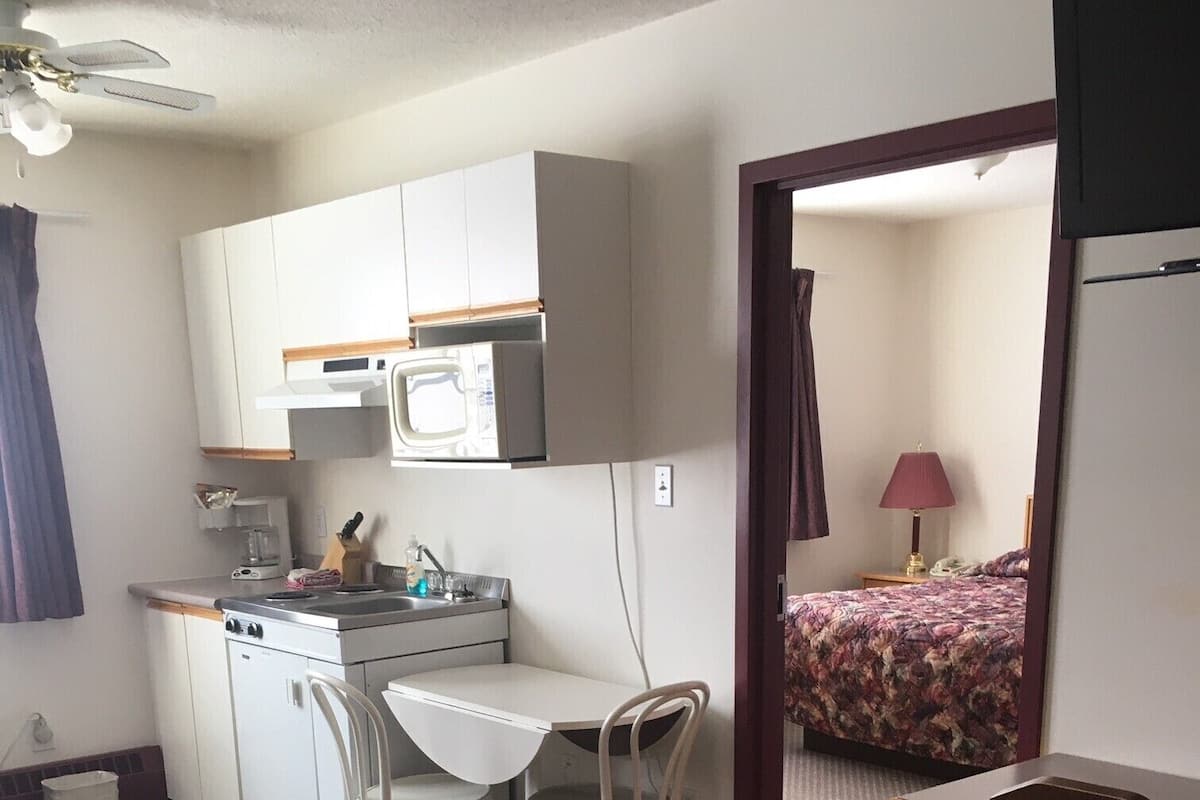 A compact hotel room with a kitchenette, a bed with a patterned bedspread, a desk, and a chair.