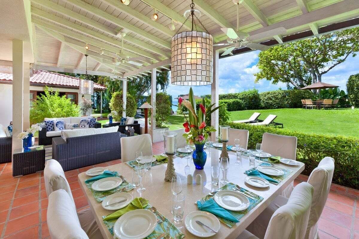 Un espace extérieur abrité, aménagé pour les repas, avec une table dressée, entouré d’une végétation luxuriante et offrant une vue sur une pelouse bien entretenue.
