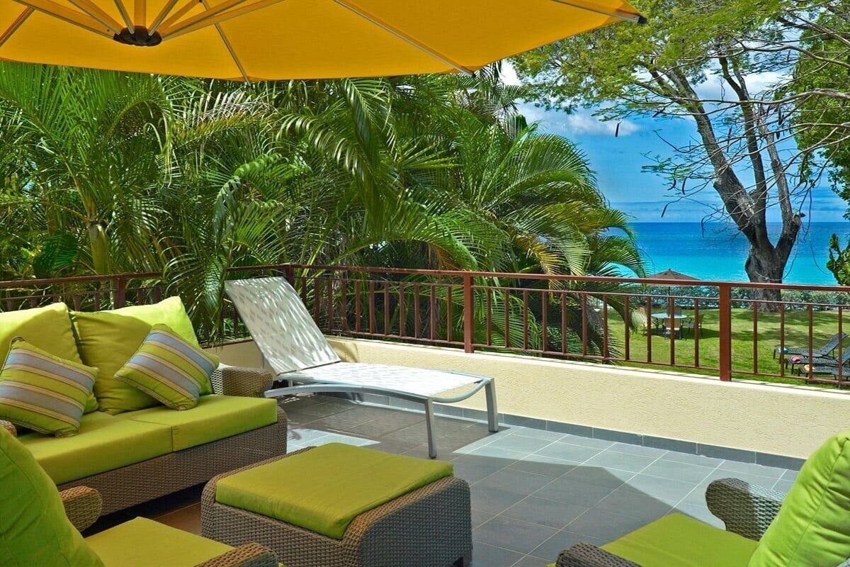 Un balcón con mobiliario de mimbre y un paraguas amarillo, con vistas a un paisaje tropical con palmeras y vista al océano.