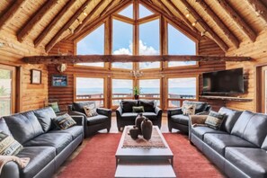 Interior de una cabaña de madera con un ventanal grande que ofrece vistas a las montañas. Hay dos sofás de cuero, una mesa de centro y un televisor montado en la pared.
