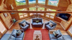 Una baita in legno con una zona salotto centrale, caratterizzata da tre divani e un tavolino da caffè, ampie finestre che offrono viste panoramiche e un lampadario.