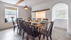 Zona pranzo con tavolo in legno apparecchiato per sei persone, circondato da sedie scure. Sopra il tavolo, un lampadario, e le pareti della stanza sono bianche con travi a vista in legno.