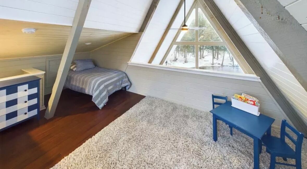 Une petite chambre mansardée avec un plafond en pente, un lit, un bureau et une fenêtre donnant sur les arbres.