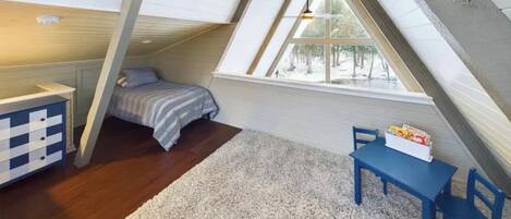Une petite chambre mansardée avec un plafond en pente, un lit, un bureau et une fenêtre donnant sur les arbres.