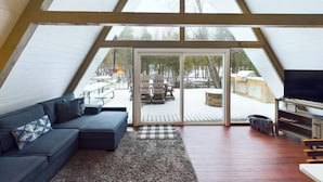 Ein gemütliches Dachzimmer mit einer dunkelgrauen Eckcouch, einem Flachbildfernseher und einer großen Schiebeglastür zu einer verschneiten Terrasse mit Gartenmöbeln.