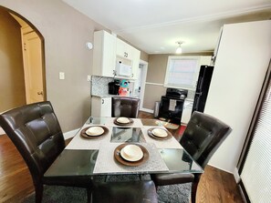 A dining area with a glass table, four chairs, and a kitchen in the background.