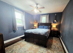 A bedroom with a bed, two bedside lamps, a ceiling fan, and a guitar on the wall.