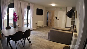 Una sala de estar moderna con un sofá, una mesa de comedor con sillas, un televisor montado en la pared y un ventanal con cortinas.