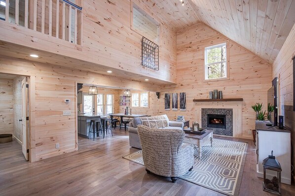 Une cabane en bois spacieuse, dotée d’un plafond haut, d’une cheminée, d’un coin repas et d’un salon confortable agrémenté d’un tapis à motifs.