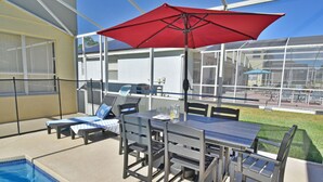 Patio esterno con piscina, tavolo e sedie, e un ombrellone rosso.