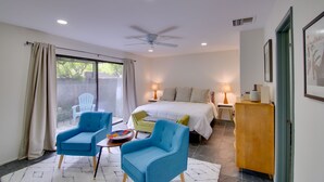 A bedroom with a bed, two blue chairs, a small table, a wooden cabinet, and a ceiling fan.