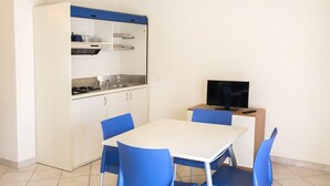 A compact kitchen with a stove, sink, and upper cabinets, a dining table with blue chairs, and a television on a small table. - M030 - Marcelli, new two-room apartment facing the sea (Marcelli)