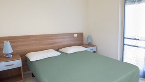 A single bed with a green bedspread, two white pillows, a wooden headboard, a nightstand with a lamp, and a window with curtains. - M030 - Marcelli, new two-room apartment facing the sea (Marcelli)