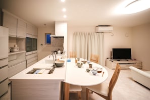 A modern kitchen with a central island, dining table, and chairs. There is a built-in oven, microwave, and a flat-screen TV in the living area.