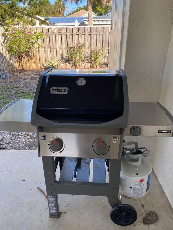 A Weber Spirit II grill with a propane tank attached, located on a concrete patio with a wooden fence and plants in the background.