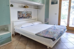 A single-bed room with a white mattress, a patterned blanket, a painting above the bed, and a view through a glass door.