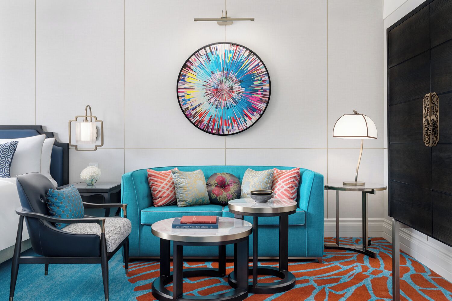 Una sala moderna con un sofá azul, dos mesas redondas, una mesa de centro, una lámpara y un cuadro abstracto de colores en la pared.
