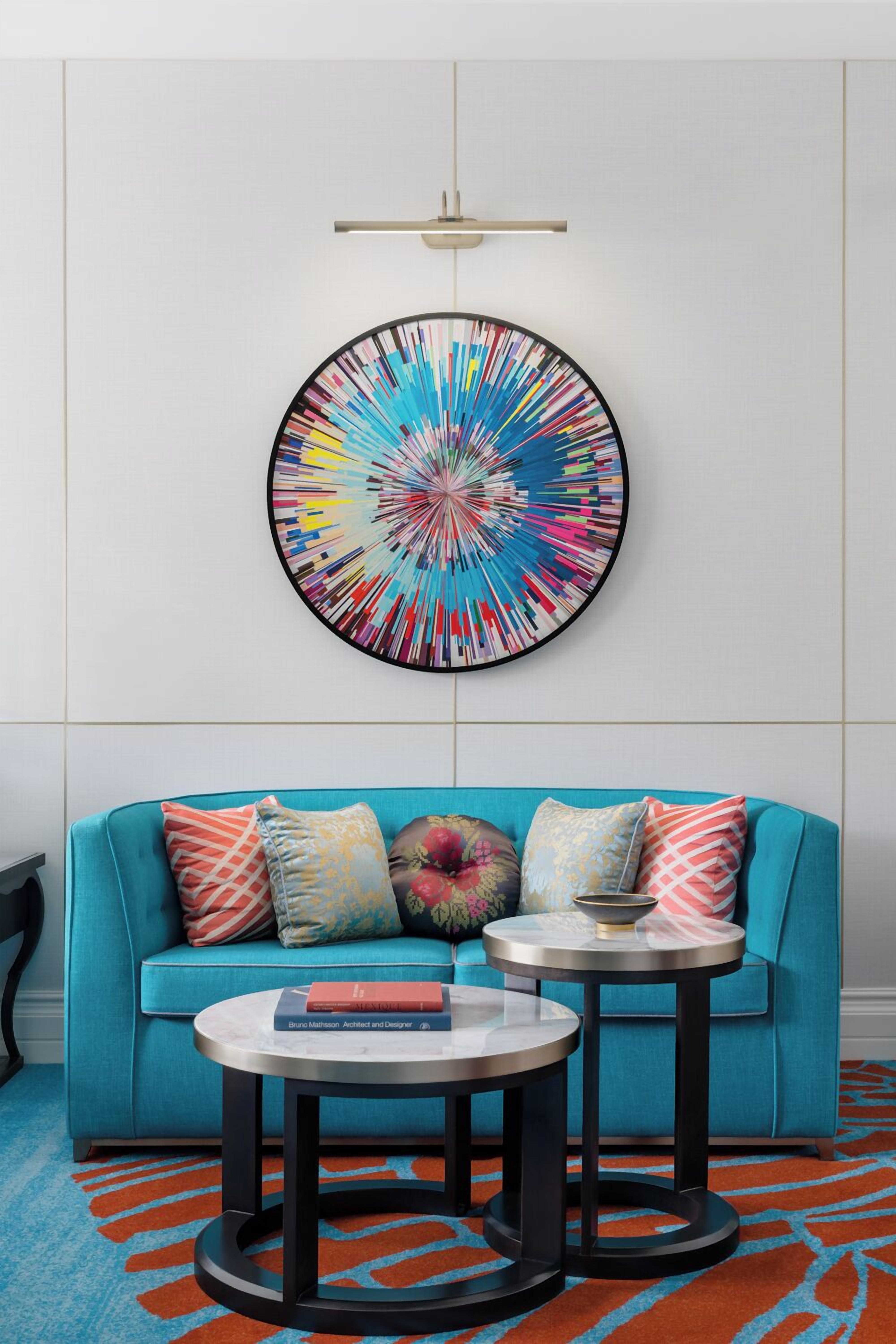 A modern living room with a teal sofa, two round tables, and a colorful abstract wall art piece.