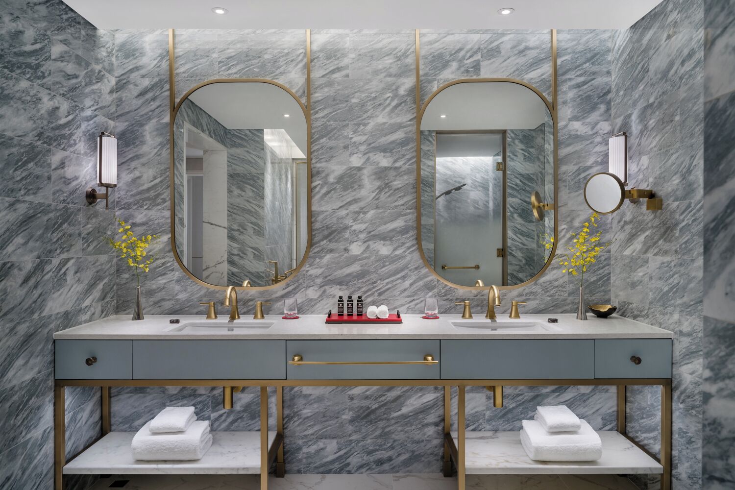 A modern bathroom with marble walls, a dual sink vanity, gold fixtures, and a large mirror.