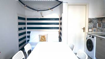 Ein kleines Zimmer mit Bett, Tisch, Stühlen, einer Waschmaschine und einem Moskitonetz an der Decke.