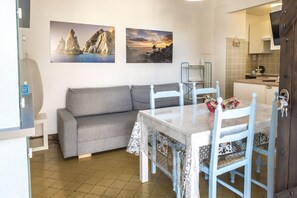 A compact living space with a sofa, a dining table with chairs, a kitchen area, and two wall-mounted pictures.