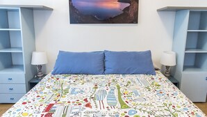 Una camera da letto con un letto, due cuscini blu, una coperta colorata, un comodino con una lampada e un quadro appeso al muro raffigurante un tramonto.