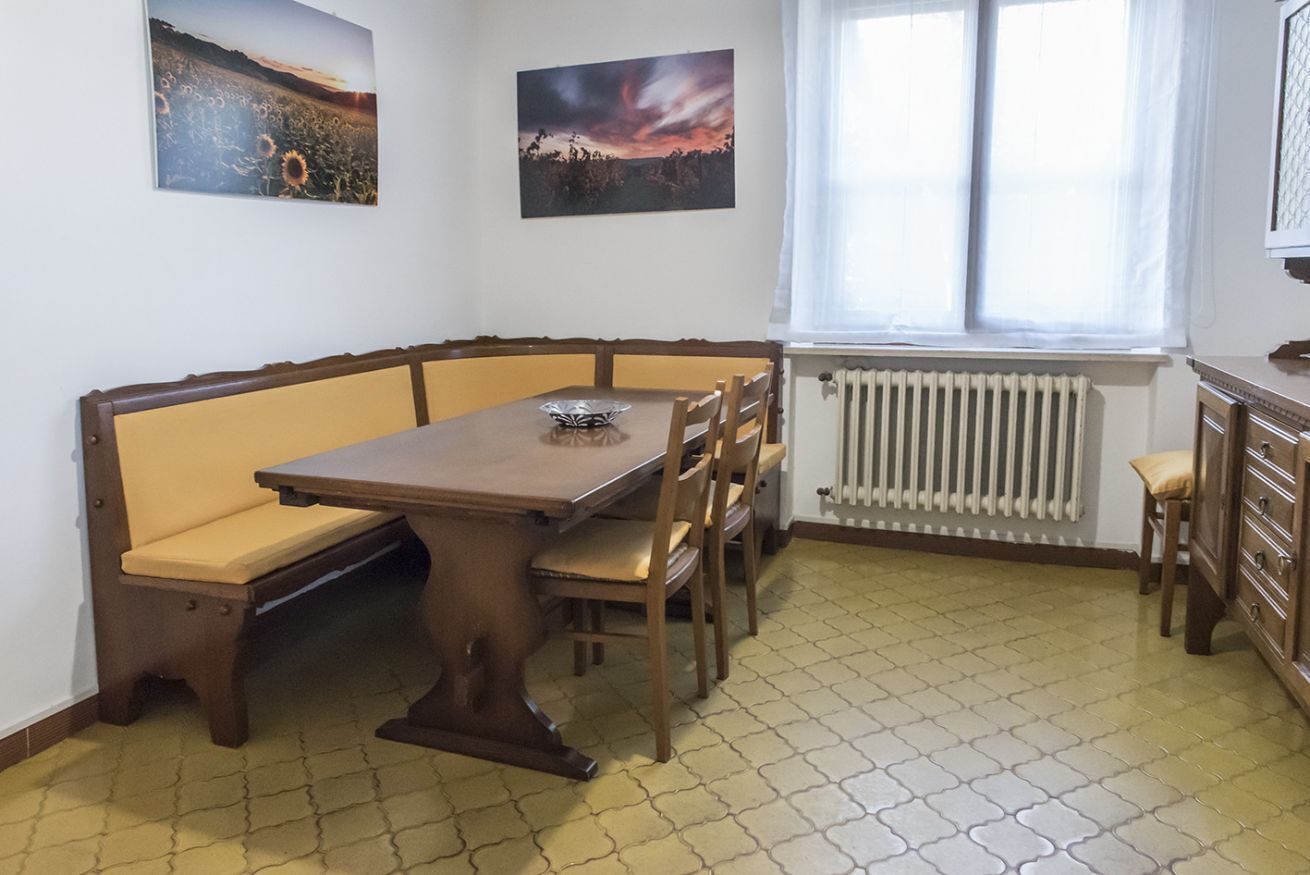 A dining area with a wooden table and bench, two wall-mounted paintings, a radiator, and a window with curtains.