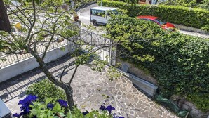 Un espace extérieur pavé de pierres, délimité par une clôture en métal, avec un arbre et une voiture rouge garée dans la rue.
