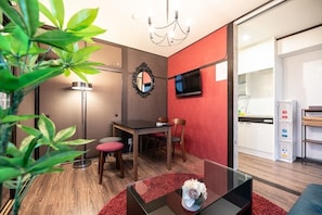 A modern living room with a dining area, a TV mounted on the wall, and a kitchen visible through an open door.