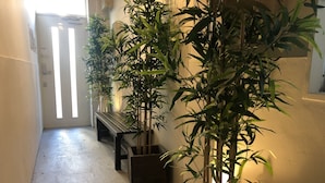 A hallway with a wooden bench, several potted bamboo plants, and a door with vertical blinds. - R-Guest Residence (Osaka)