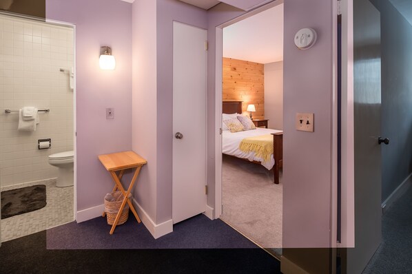 A hotel room with a bathroom, a bed with a wooden headboard, and a small table with a basket. - Classic Stowe Ski 3 Bedroom Chalet by RedAwning (Stowe)
