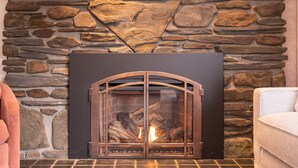 A stone fireplace with a glass door, a fire burning inside, and a cushioned armchair to the right. - Classic Stowe Ski 3 Bedroom Chalet by RedAwning (Stowe)