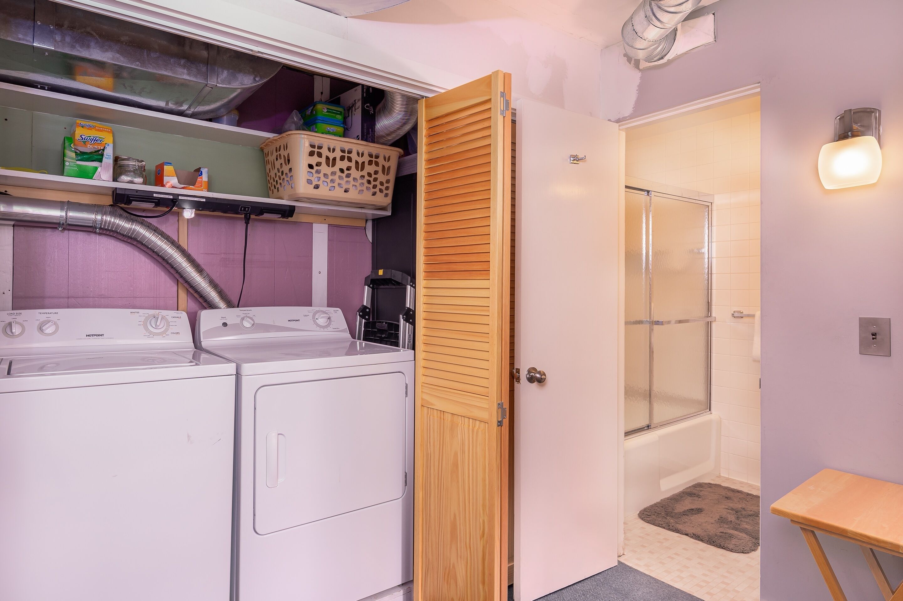 A laundry area with two white washing machines, a shelf with cleaning supplies, and a wooden door leading to a shower.