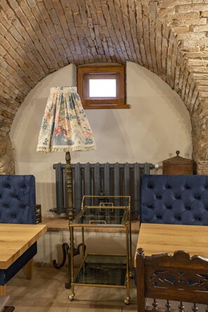 Une pièce avec un plafond en briques, une petite fenêtre, un radiateur, une table en bois, une chaise recouverte de tissu bleu et une vitrine.