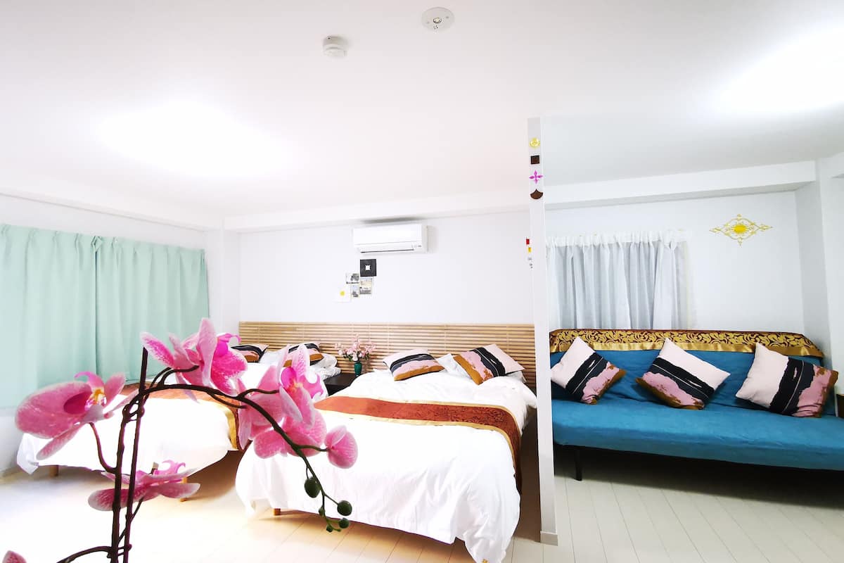 Quarto de hotel com duas camas, uma com colcha branca e outra com colcha azul. Há uma janela com cortinas claras, um aparelho de ar condicionado na parede e um ornamento decorativo.