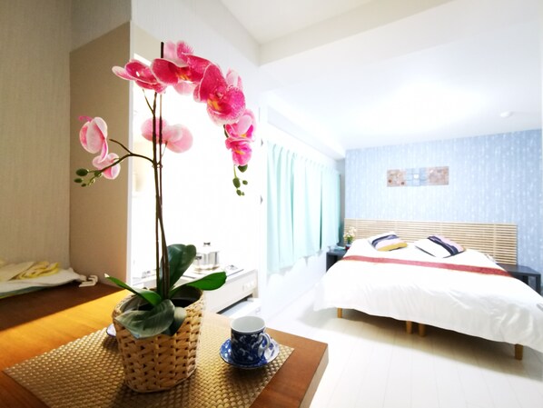 A hotel room with a bed, a bedside table with a cup and saucer, a potted orchid, and a view of another room.