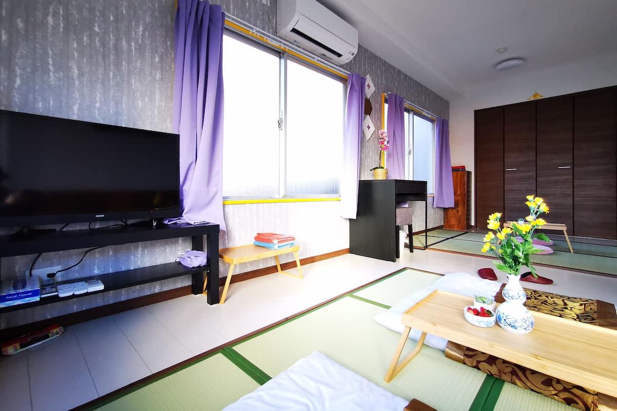 Uma sala de estar moderna com uma televisão de tela plana, uma mesa pequena com um vaso de flores, esteiras de tatame e uma porta deslizante com cortinas roxas.